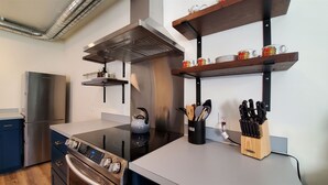 Una cocina moderna con electrodomésticos de acero inoxidable, una placa de inducción negra y estantes de madera con tazas y utensilios.