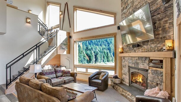 Un salón amplio con chimenea de piedra, un ventanal grande con vistas a un bosque y una escalera con barandilla de metal.