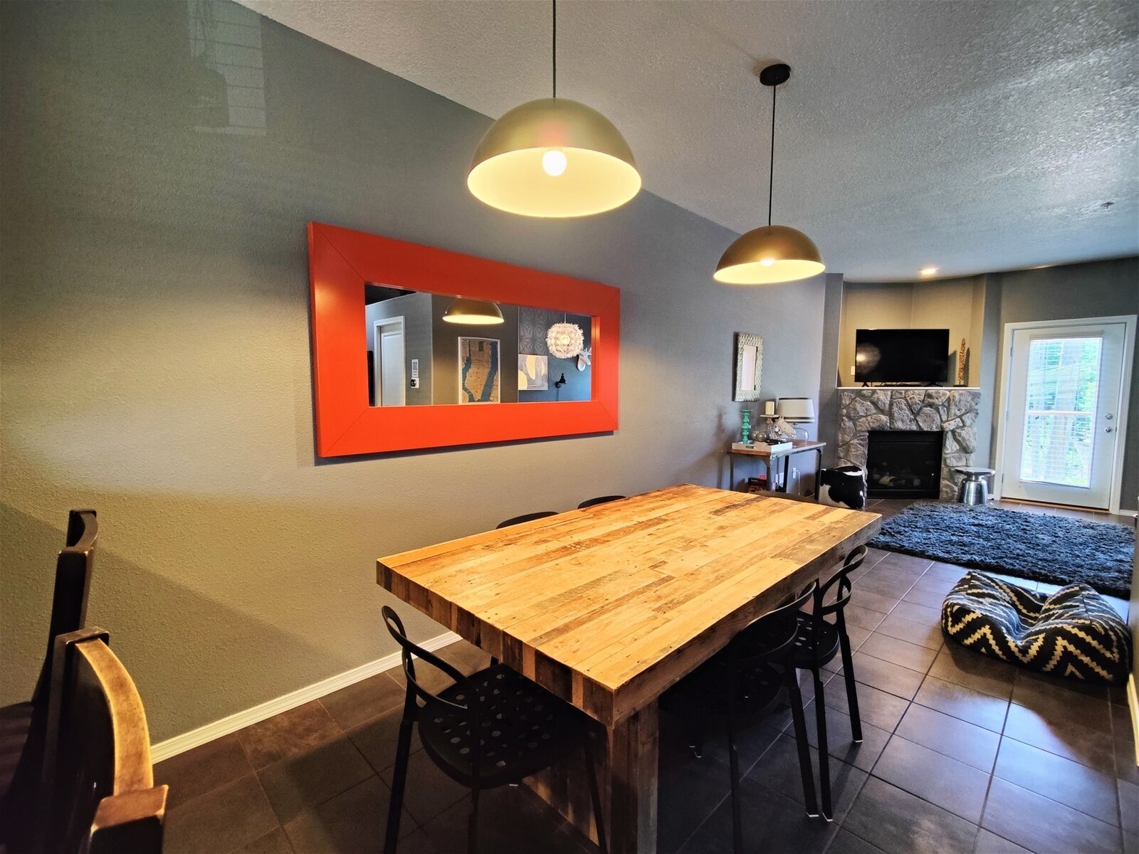 Un coin repas avec une table et des chaises en bois, deux lampes suspendues, un miroir à cadre rouge, une cheminée et un téléviseur fixé au mur.