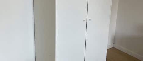 A white wardrobe with two doors and a flat base, positioned against a wall with a wooden floor.