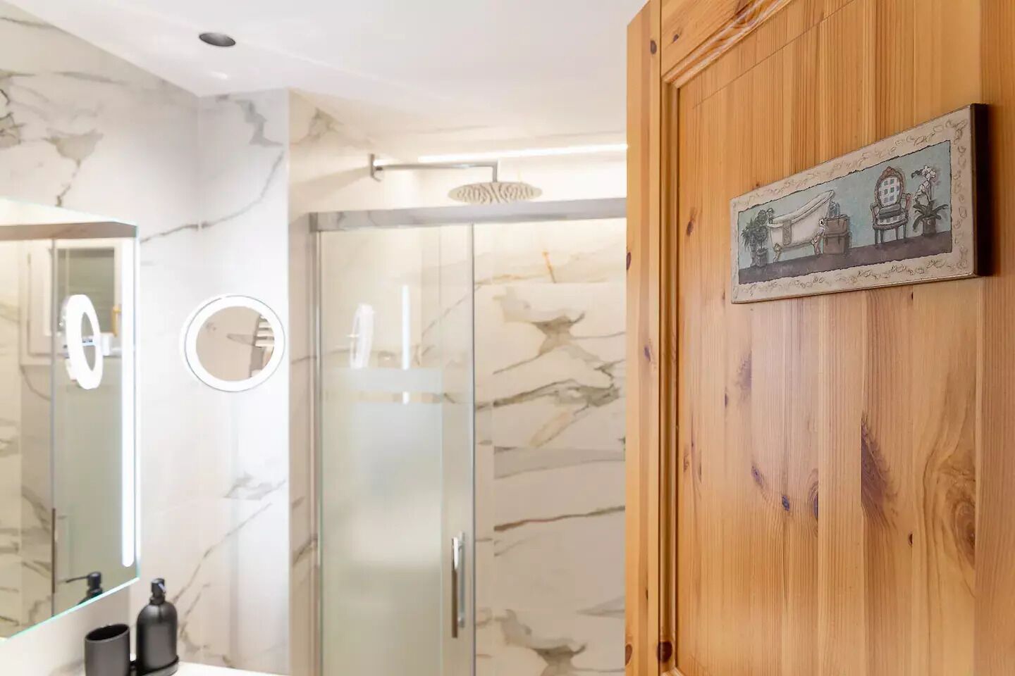 A bathroom with a marble wall, a wooden door, and a mirror with a light.