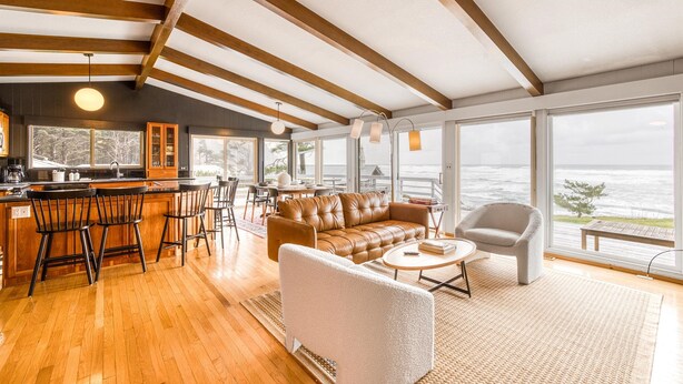 A spacious living area with a wooden ceiling, a leather sofa, a white armchair, and a bar area with stools. - Ocean Views | Arch Cape - OPEN Sept 7 - 13! (Arch Cape)