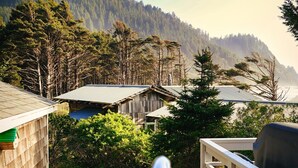 A view of a forested hillside with a cabin and a metal roof structure in the foreground. - Ocean Views | Arch Cape - OPEN Sept 7 - 13! (Arch Cape)