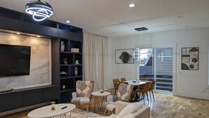 A modern living room with a large flat-screen TV, a dark bookshelf, a dining area with a table and chairs, and a view through a glass door. - Stunning Home Getaway
Modern house with a rooftop / 6 minute from Broadway (Nashville)