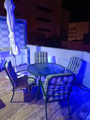 An outdoor patio area with a round table and four striped chairs, a white umbrella, and a tiled floor.