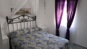 A bedroom with a patterned bedspread, a white bedside table, a nightstand, a decorative wall hanging, and purple curtains. - Casa Familiar (San Fernando)