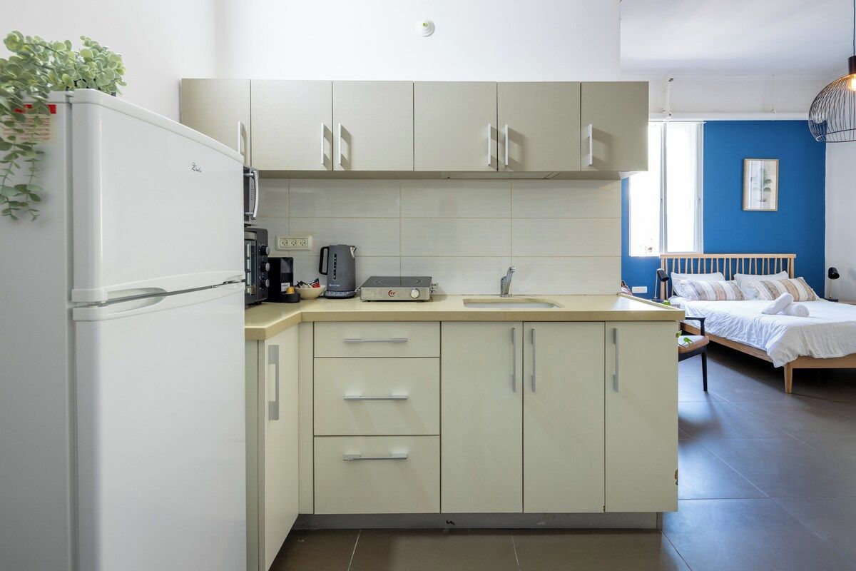 A compact kitchen with a refrigerator, microwave, and coffee maker. A bed with a wooden headboard is visible in the adjacent room.