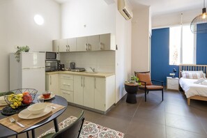 Una cocina compacta con microondas, tostadora y lavamanos, un comedor con mesa y sillas, y un dormitorio con cama y mesita de noche.