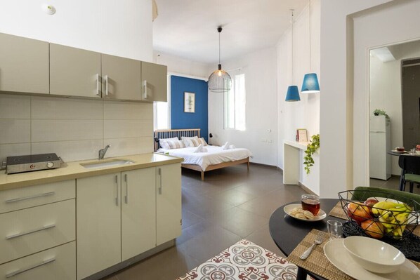 Habitación de hotel compacta con kitchenette, una cama con ropa de cama blanca, una zona de comedor con mesa y sillas, y una pared azul de acento.
