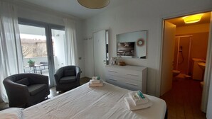 Une chambre d’hôtel avec un lit, deux fauteuils, un bureau, un miroir et une salle de bain visible à travers une porte ouverte.