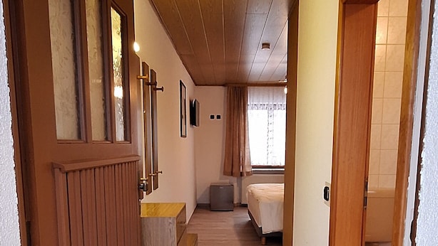 A hallway with wooden paneling, a radiator, and a tiled floor leading to a bedroom with a bed and a window. - Sharmas (Ilmenau)