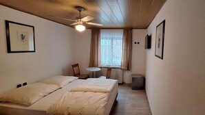 A bedroom with a bed, a ceiling fan, a window with curtains, a small table, and two chairs. - Sharmas (Ilmenau)