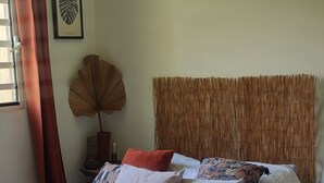 Un lit bien fait, avec une couvre-lit à rayures, un éventail décoratif et une plante encadrée accrochée au mur.