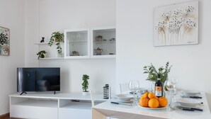 Una sala de estar moderna con un televisor de pantalla plana, estanterías blancas, una mesa de comedor preparada con platos, vasos y una botella de vino, y un cuadro en la pared.
