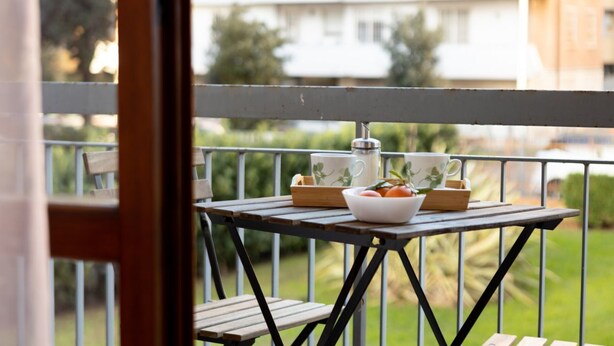 Un balcón con una mesa dispuesta para dos personas, que incluye una taza blanca, un termo de acero inoxidable y un bol con tomate.