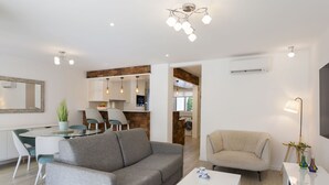 Una sala de estar moderna con un sofá gris, una mesa de centro blanca y un sillón color beige. Hay una zona de comedor con una mesa y sillas de vidrio, y una cocina con detalles de madera.
