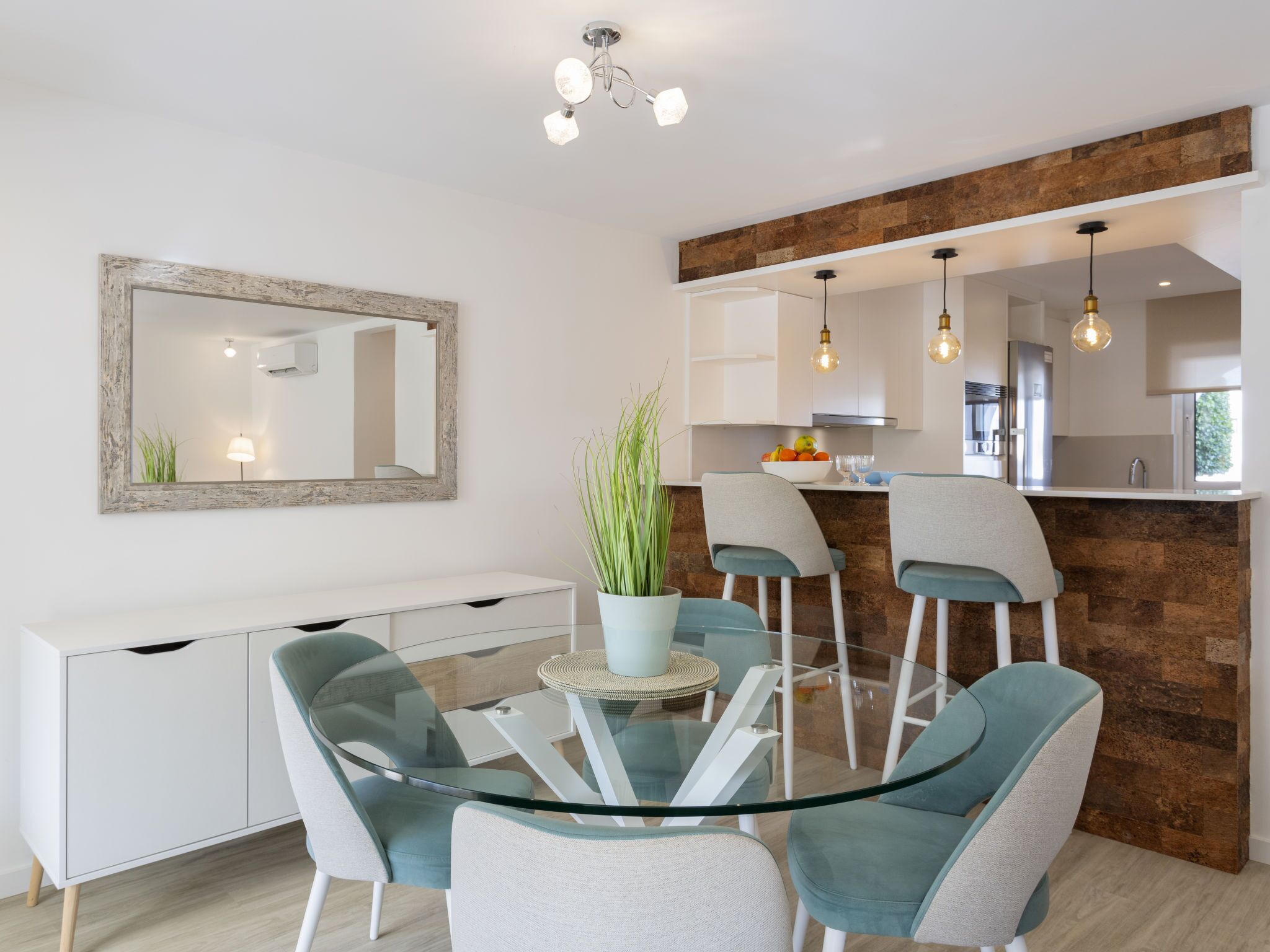 Una cocina moderna con una pared de acento de piedra, un área de comedor con mesa de vidrio y sillas color turquesa, y un mueble bajo blanco con cajones.