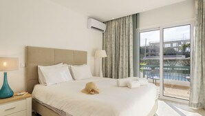 Habitación de hotel con una cama grande, una mesita de noche con lámpara, una ventana con vistas a una piscina y edificios, y un equipo de aire acondicionado en la pared.