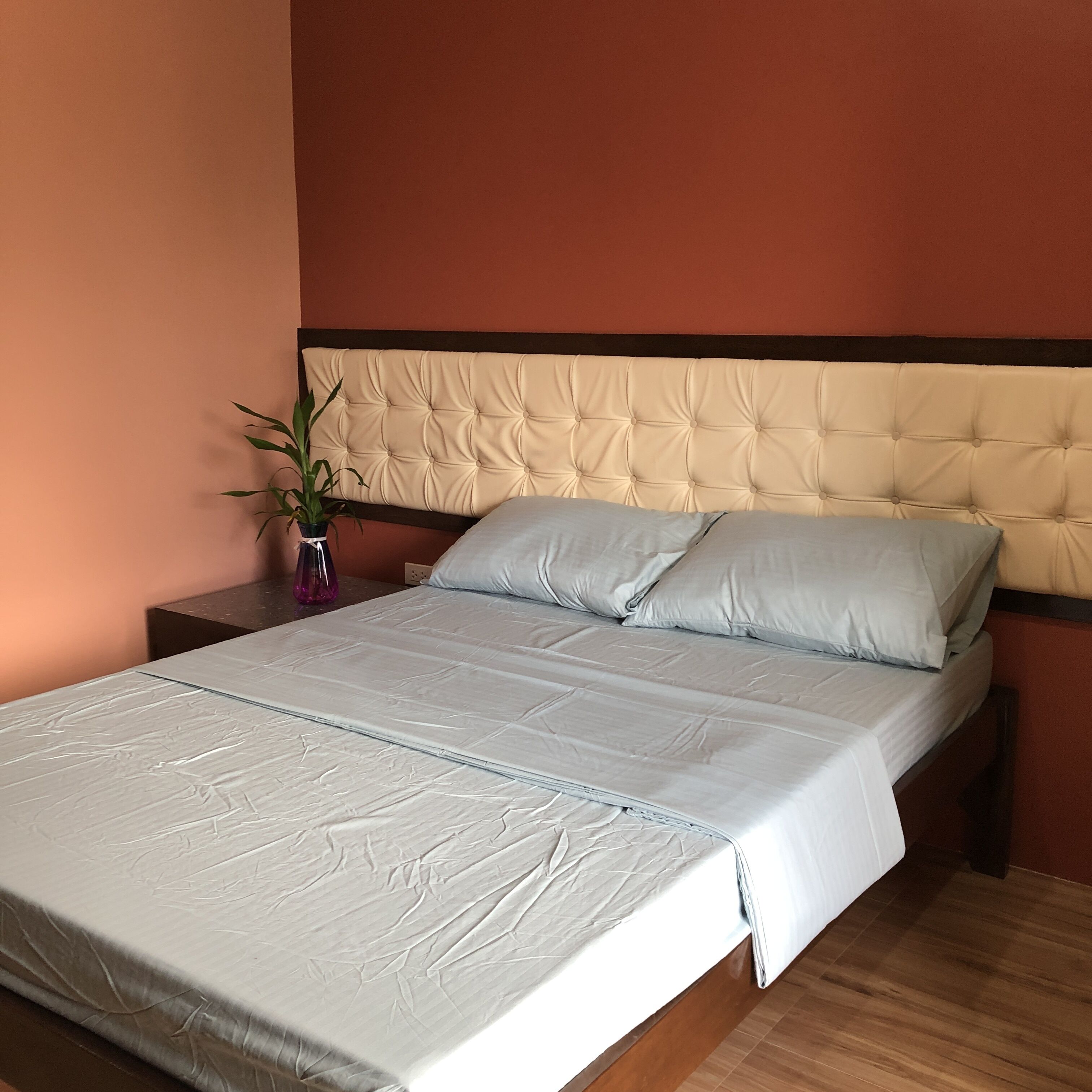Ein ordentlich bezogenes Bett mit einer geblatteten Kopfstütze, ein Nachttisch mit einer Blumenvase und ein Holzboden.