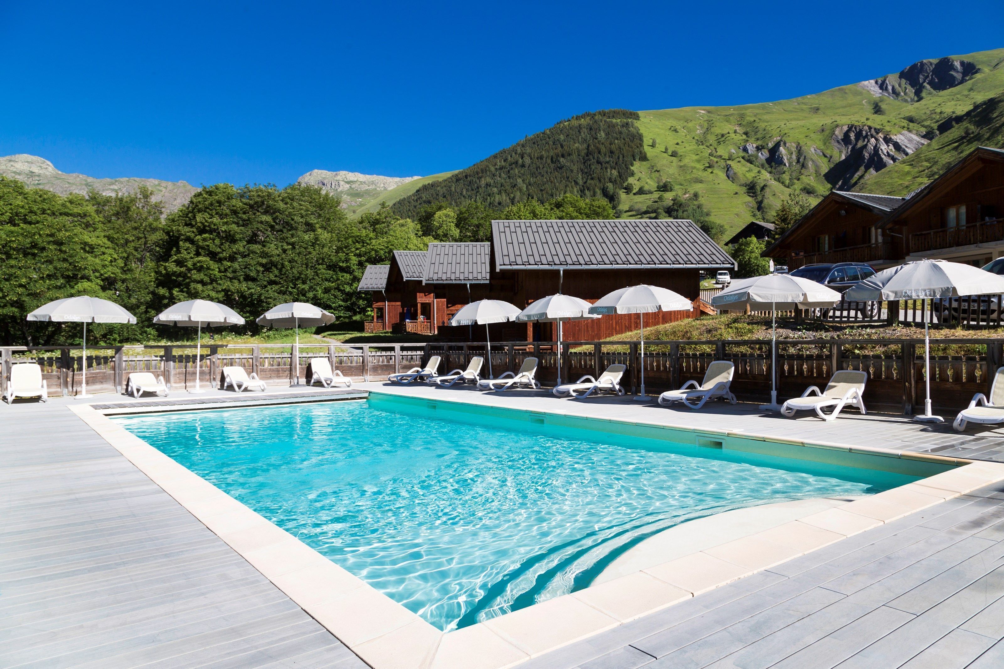 Una piscina con sillas de descanso y sombrillas, rodeada de cabañas de madera con montañas al fondo.