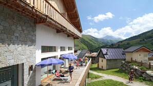 Une terrasse en bois avec des chaises longues et des parasols, un bâtiment en pierre doté d’un balcon, et une vue sur des collines verdoyantes et des montagnes en arrière-plan.