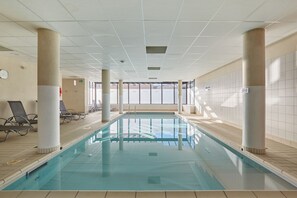 Uma piscina interior com paredes de vidro transparente, iluminada por luz natural, rodeada por espreguiçadeiras e colunas.
