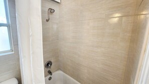 Ein Badezimmer mit beigen Fliesen, einer Dusche mit Duschhandstück und einer Badewanne mit Wasserhahn.