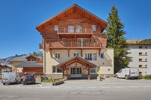 A two-story wooden building with a balcony, a sign reading 'Residence L'Ours Blanc,' and a parking area with vehicles. - Appartement avec Accès Piscine + Casiers à Ski (Les Deux Alpes)