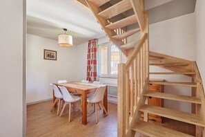A dining area with a wooden table and chairs, a staircase, and a window with curtains. - Appartement avec Accès Piscine + Casiers à Ski (Les Deux Alpes)