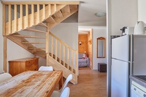 A wooden staircase leading to an upper level, a dining area with a wooden table and chairs, and a kitchen with a refrigerator.