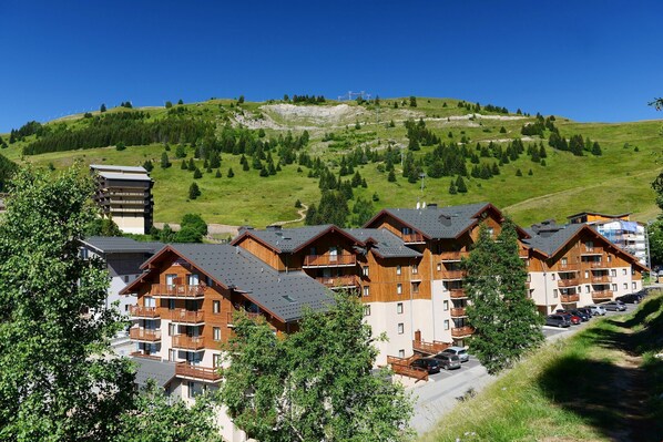 Una hilera de edificios tipo cabaña de madera con balcones, frente a un paisaje de colina verde y cielo azul despejado.