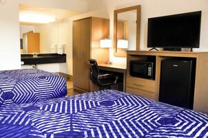A hotel room with two beds, a desk with a lamp, a television, a microwave, and a refrigerator.