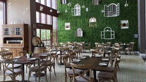 A restaurant with multiple tables and chairs, a wooden cabinet with wine bottles, and a green wall with decorative windows.