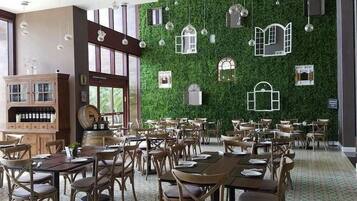 Un restaurante con varias mesas y sillas, un aparador de madera con botellas de vino y una pared verde con ventanas decorativas.