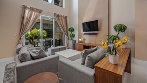A modern living room with a sofa, coffee table, and a wooden side table with yellow flowers. A flat-screen TV is mounted on the wall, and there is a balcony with a view of greenery.