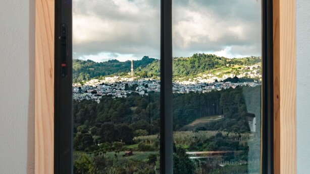 Une fenêtre dotée d’un cadre en bois et de vitres noires, qui offre une vue sur une ville avec ses bâtiments et sa végétation.