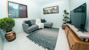 Un salon moderne avec un canapé gris, un téléviseur à écran plat, une grande fenêtre et un tableau représentant un paysage urbain, encadré.