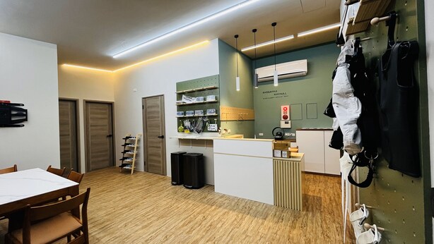 A compact room with a wooden floor, a white table with chairs, a shoe rack, a small trash bin, and a kitchenette area with a fridge, microwave, and a hanging rack with bags. - Ocean Tale Inn (Liuqiu)
