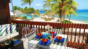 Un balcon avec une table dressée pour quatre personnes, donnant sur une plage bordée de palmiers et une piscine.