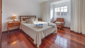 Un dormitorio ordenado con una cama de madera, una mesita de noche con lámpara, una silla y una ventana con cortinas.