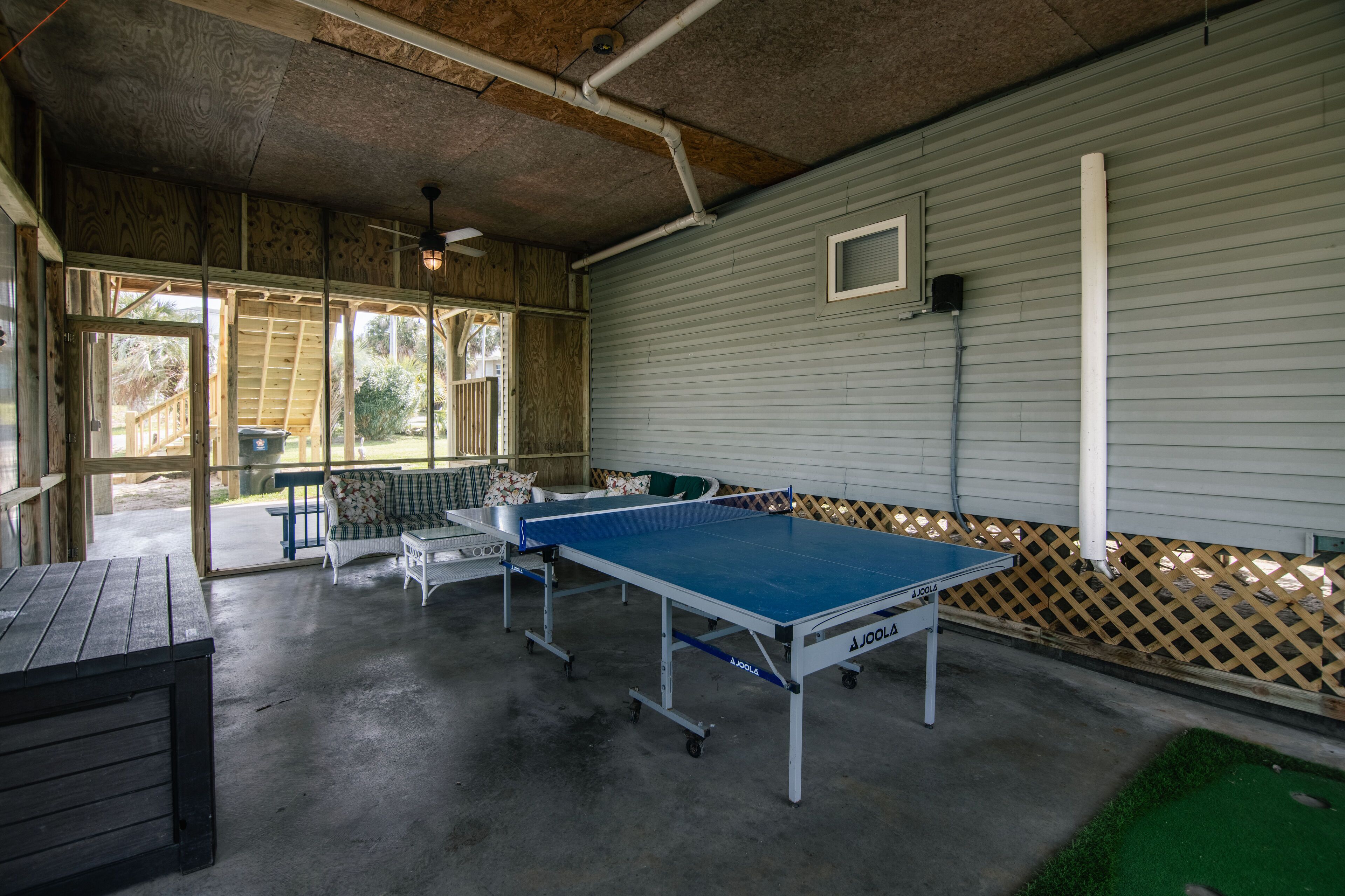 Mesa de ping pong en un espacio exterior cubierto, con un banco de madera y una pequeña mesa.