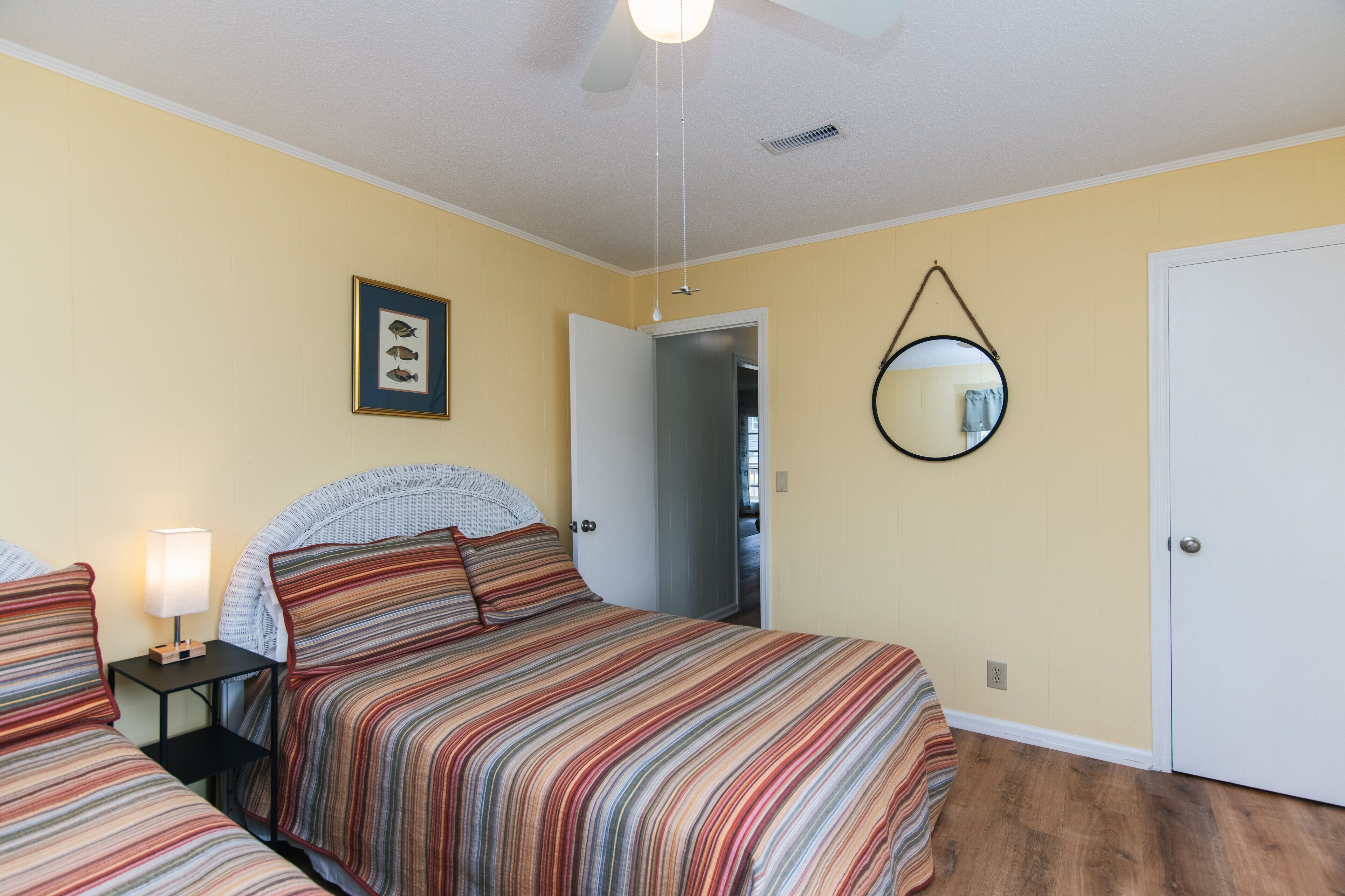 Quarto com duas camas, uma mesinha de cabeceira com abajur, um quadro na parede e uma porta que dá para outro cômodo.