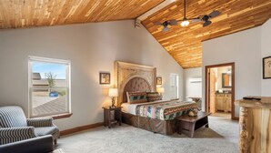 Un dormitorio con techo de madera, una cama con cabecera capitonada, sillas a rayas, una mesita y vistas al exterior a través de una ventana.