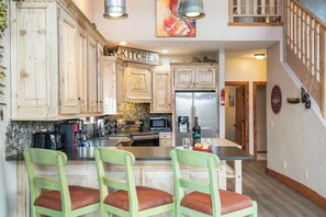 A kitchen with wooden cabinets, a stainless steel refrigerator, a microwave, and a coffee maker. There are three green chairs with red cushions. - On Table Rock Shoreline w/ Lake Views, Private Hot Tub, Billiards, Boat Slip & Cmty Amenities (Branson)