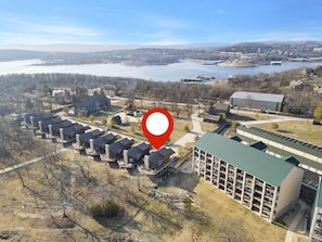 An aerial view of a residential area with detached houses, a large building with a green roof, and a body of water in the background. - On Table Rock Shoreline w/ Lake Views, Private Hot Tub, Billiards, Boat Slip & Cmty Amenities (Branson)