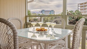 Une table à plateau en verre, sur laquelle sont disposés six verres à margarita et un bol de chips, entourée de chaises en osier, dans un espace extérieur abrité offrant une vue sur les bâtiments et les palmiers.