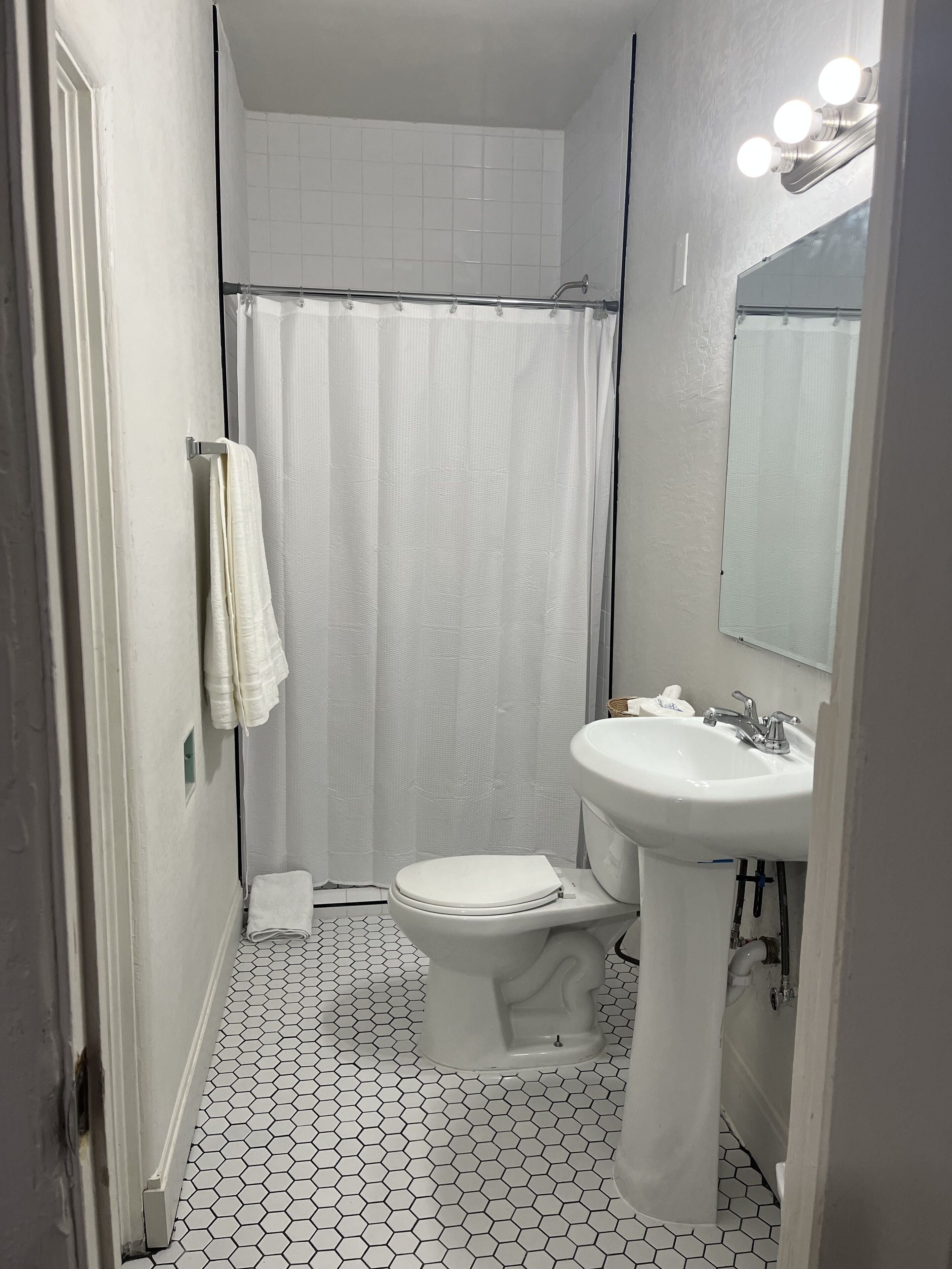 Un baño con inodoro y lavamanos blancos, cortina de ducha y espejo con iluminación.