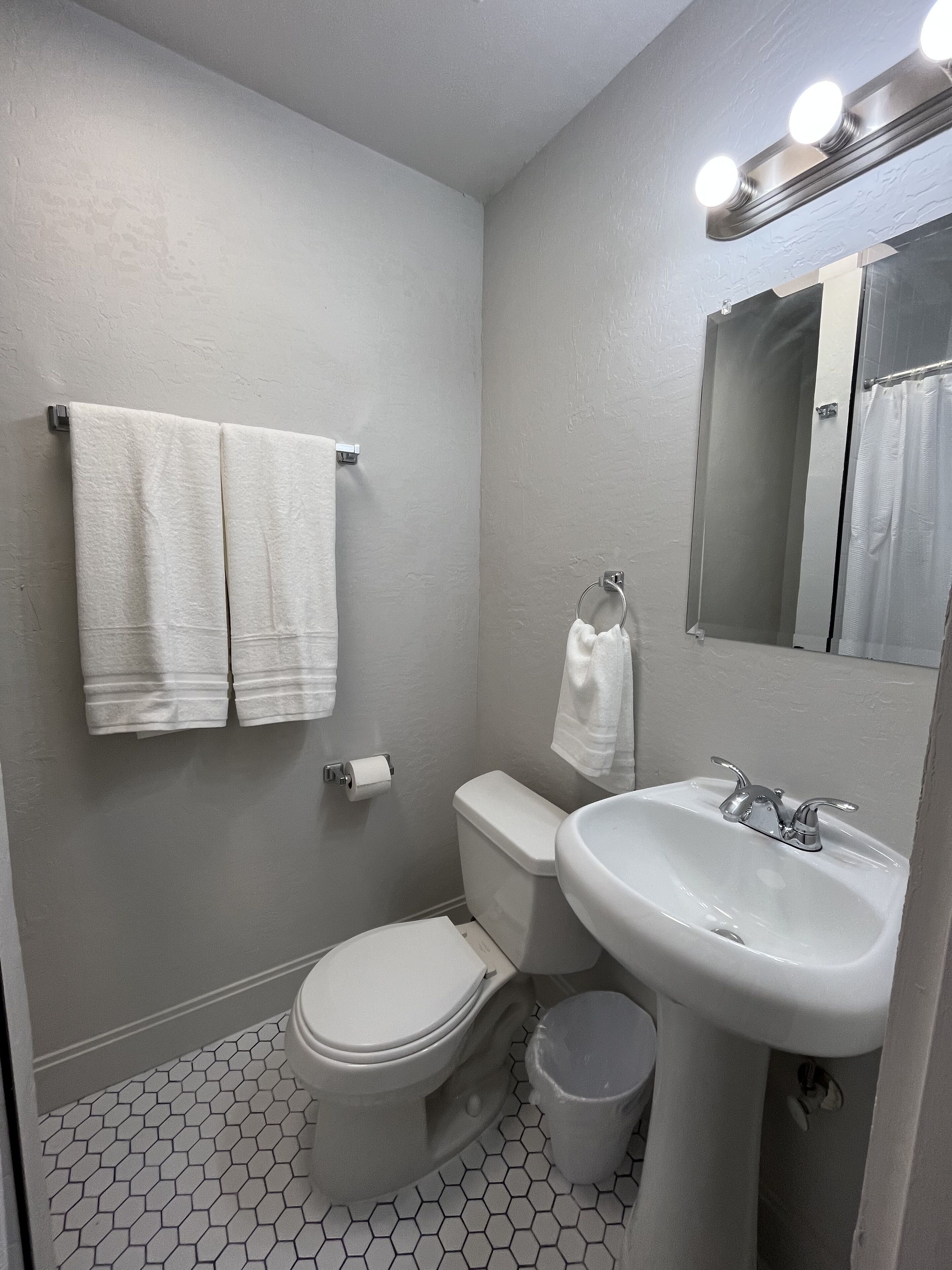 Une petite salle de bain propre, équipée de toilettes, d’un lavabo et d’un miroir. Deux serviettes blanches sont accrochées au mur, et il y a un rouleau de papier toilette. Le sol est recouvert de carreaux hexagonaux.