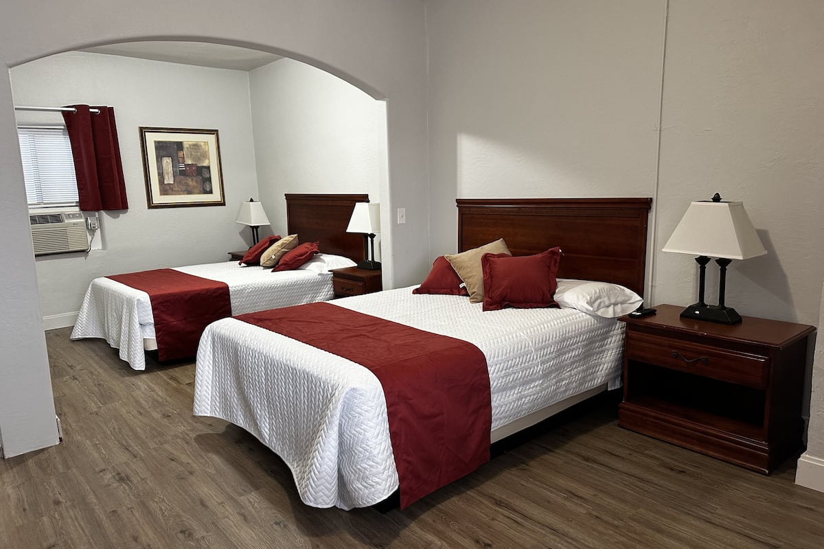 A hotel room with two beds, each with a wooden headboard, white bedding, and red throw pillows. There is a nightstand with a lamp, a framed picture on the wall, and a window with blinds.