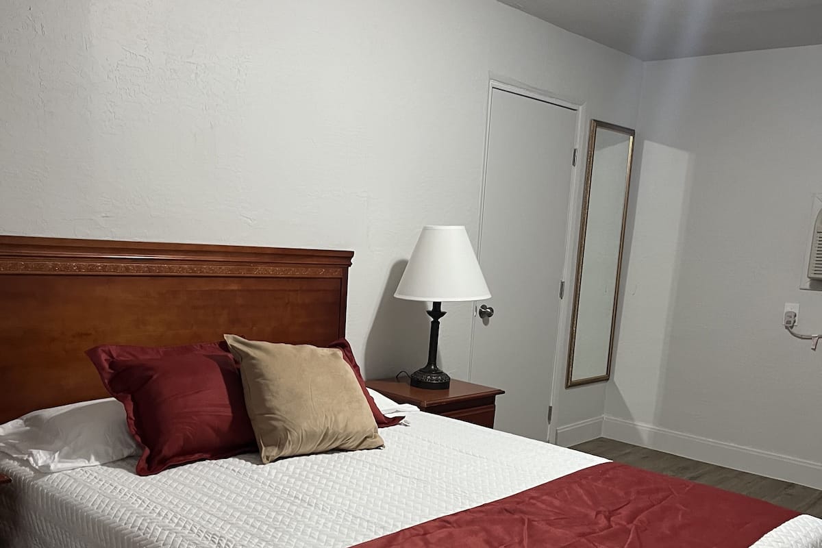 A neatly made bed with a wooden headboard, a red and white bedspread, and two pillows. There is a bedside table with a lamp and a mirror. The room has white walls and a wooden floor.